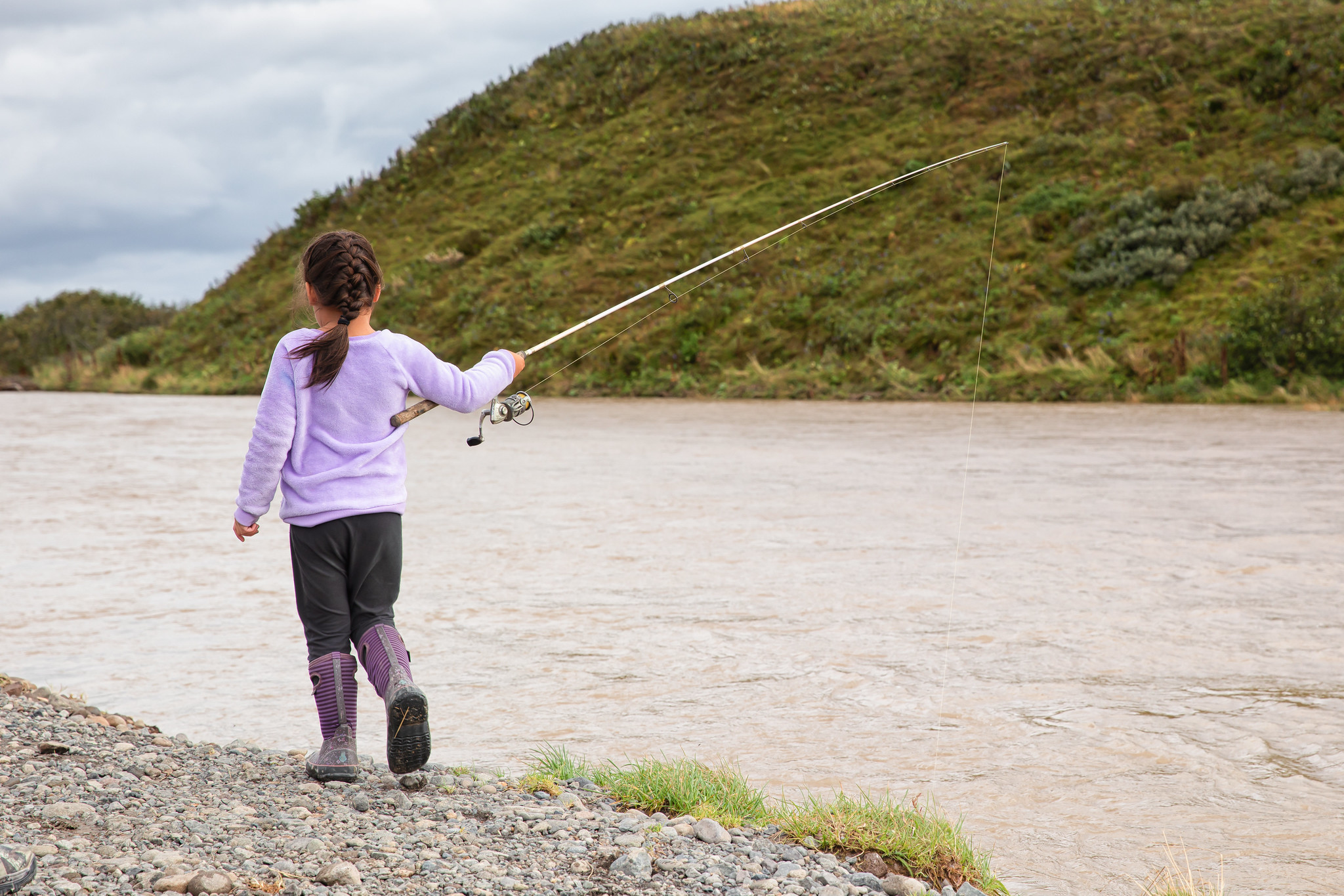 A girl fishes along the bank of a brown river with a green hillside on the opposite bank.