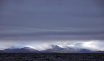 Dark clouds settle over mountains with a gray sea in front. 