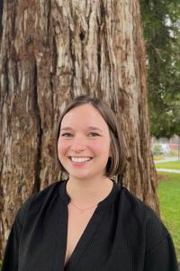 A young woman, smiling, stands in front of a tree.